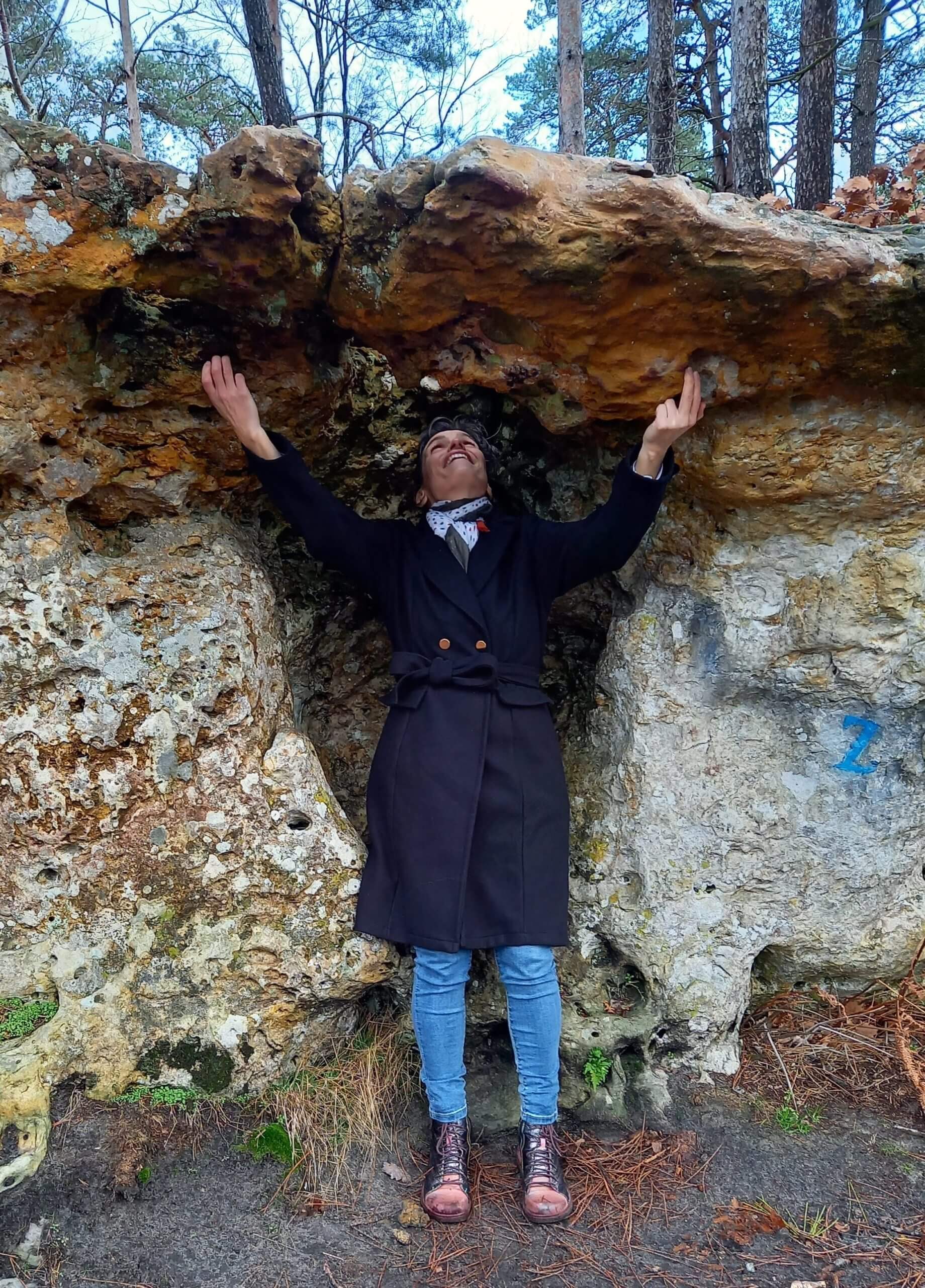 Jeune femme debout avec un manteau noir au sein d'un rocher avec un trou au-dessus d'elle où elle voit le ciel au sein d'une forêt.